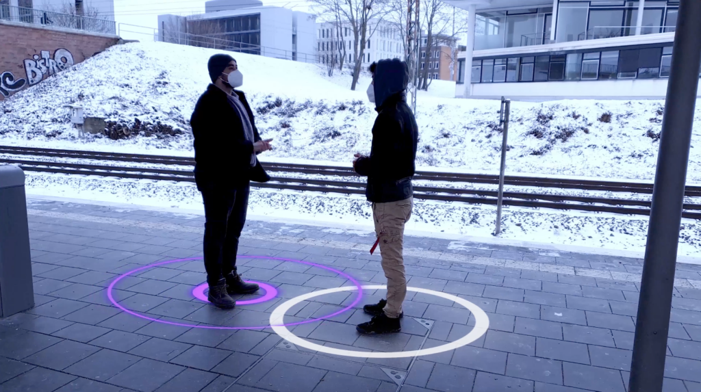 Image of 2 persons with masks talking at the trainstation. One has purple circles underneath himself and the other a white circle.