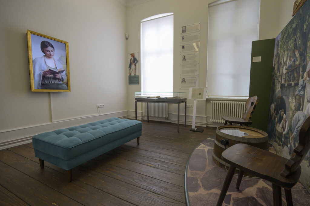 Picture of a young woman reading a book inside of a golden picture frame. The scene outside shows a room with a blue couch, wooden chairs around a halfed barrel and a glass showcase.